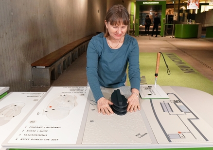 Eine Frau mit Blindenstock steht an einer tastbaren Übersichtskarte im Museum